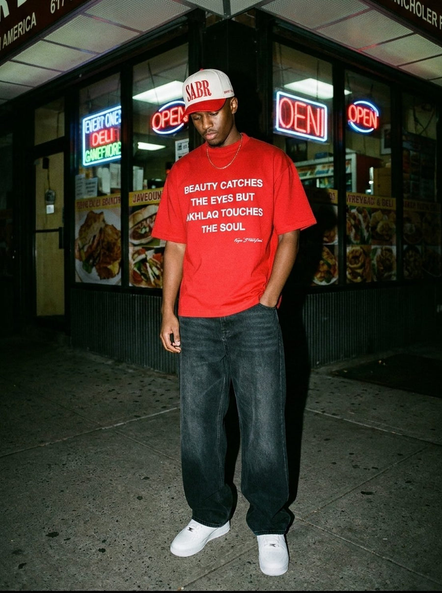 Ivory and red Sabr cap worn with the red Akhlaq Over Beauty tee outside a New York deli