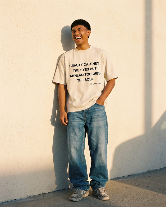Akhlaq Over Beauty natural boxy tee against a warm plaster wall