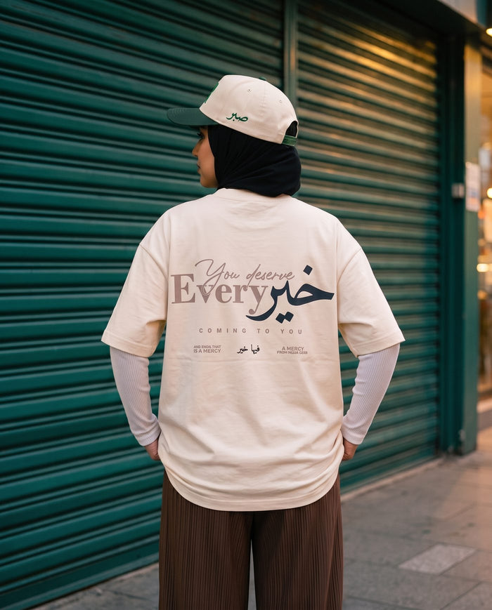 You Deserve Every Khayr natural boxy tee back print in front of a green roller shutter at dusk