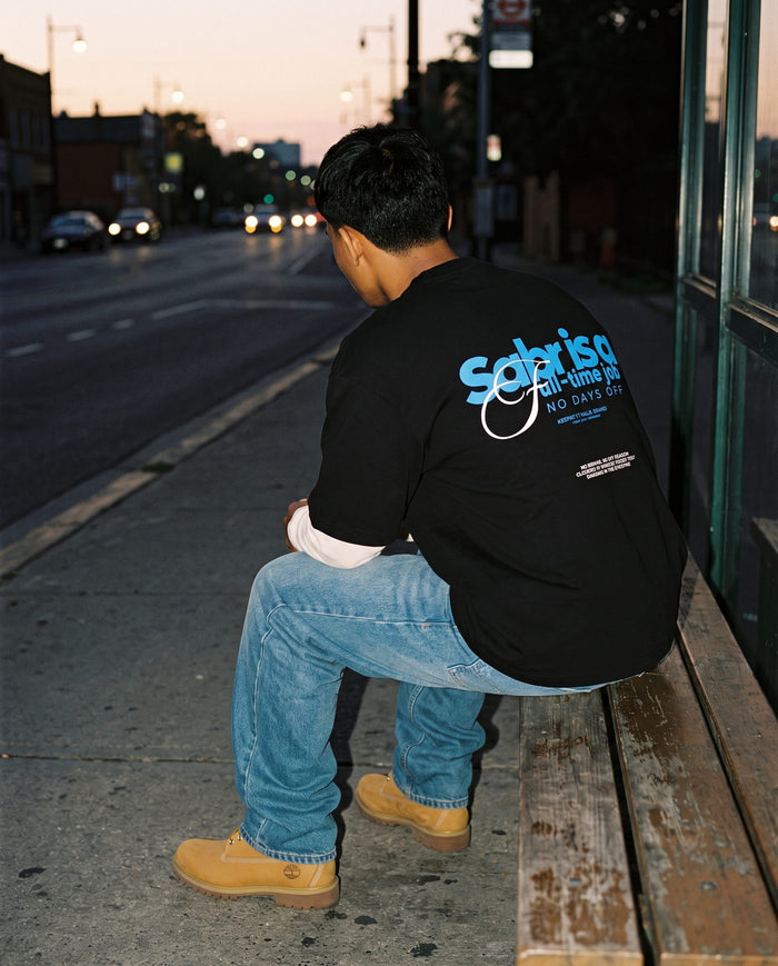 Full-Time Sabr black boxy tee seen from behind, seated kerbside at dusk
