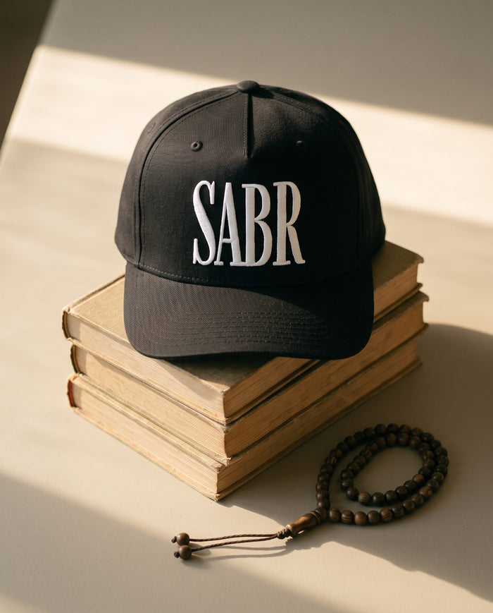 All-black Sabr Cap with white embroidered lettering, resting on a stack of vintage books beside a wooden misbaha