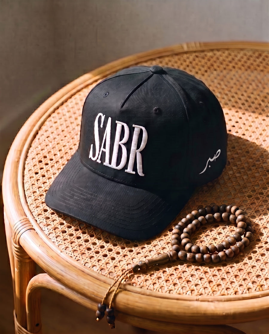 All-black Sabr Cap with white embroidered lettering, on a cane table beside wooden prayer beads