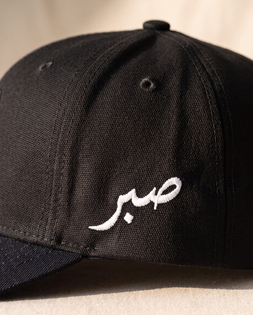 Close detail of the Arabic word sabr embroidered in white on the left side panel of the all-black Sabr Cap