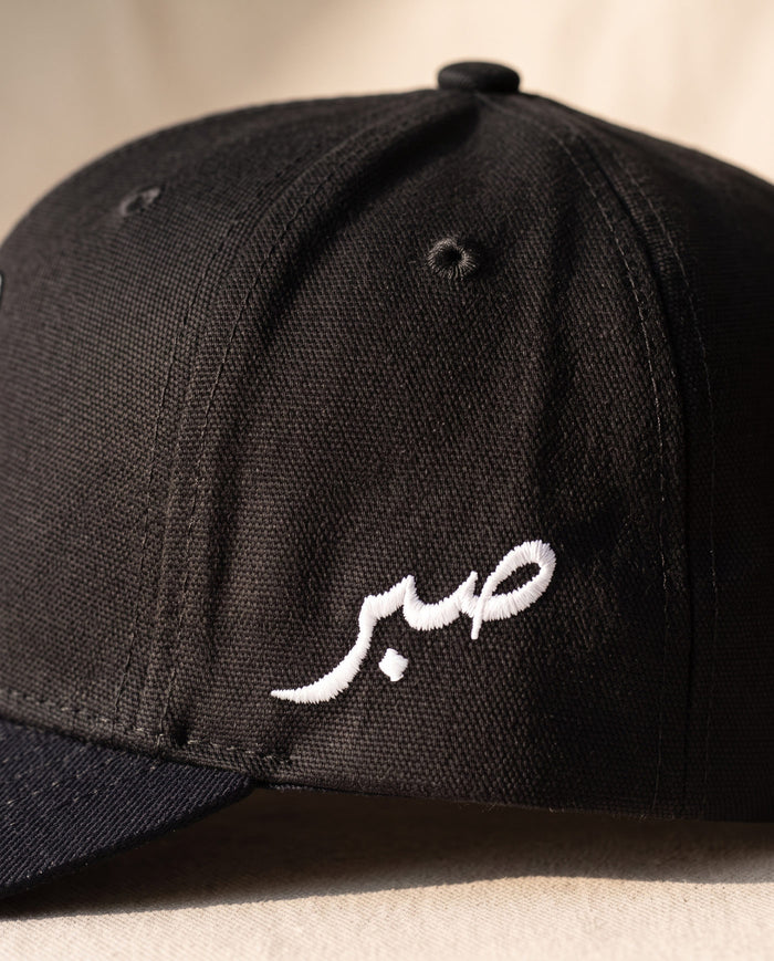 Close detail of the Arabic word sabr embroidered in white on the left side panel of the all-black Sabr Cap