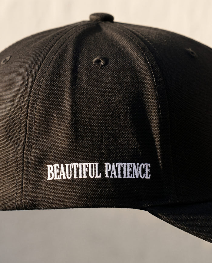 Close detail of BEAUTIFUL PATIENCE embroidered in white on the right side panel of the all-black Sabr Cap