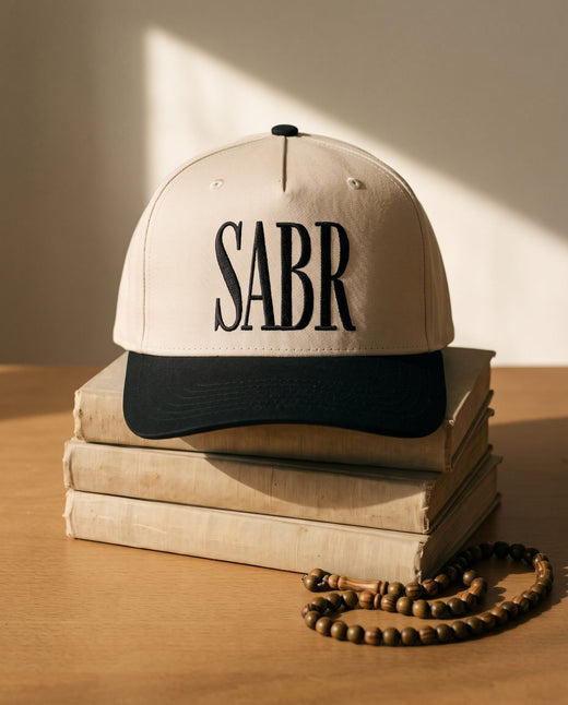 Sabr Cap in ivory with a black brim and black embroidered lettering, resting on a stack of vintage books beside a wooden misbaha