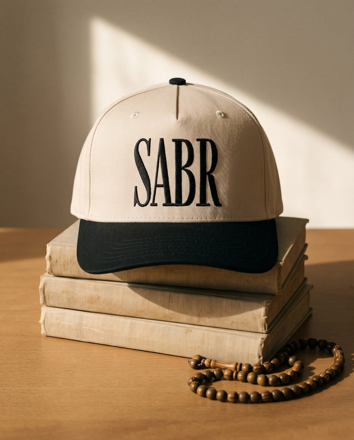 Sabr Cap in ivory with a black brim and black embroidered lettering, resting on a stack of vintage books beside a wooden misbaha