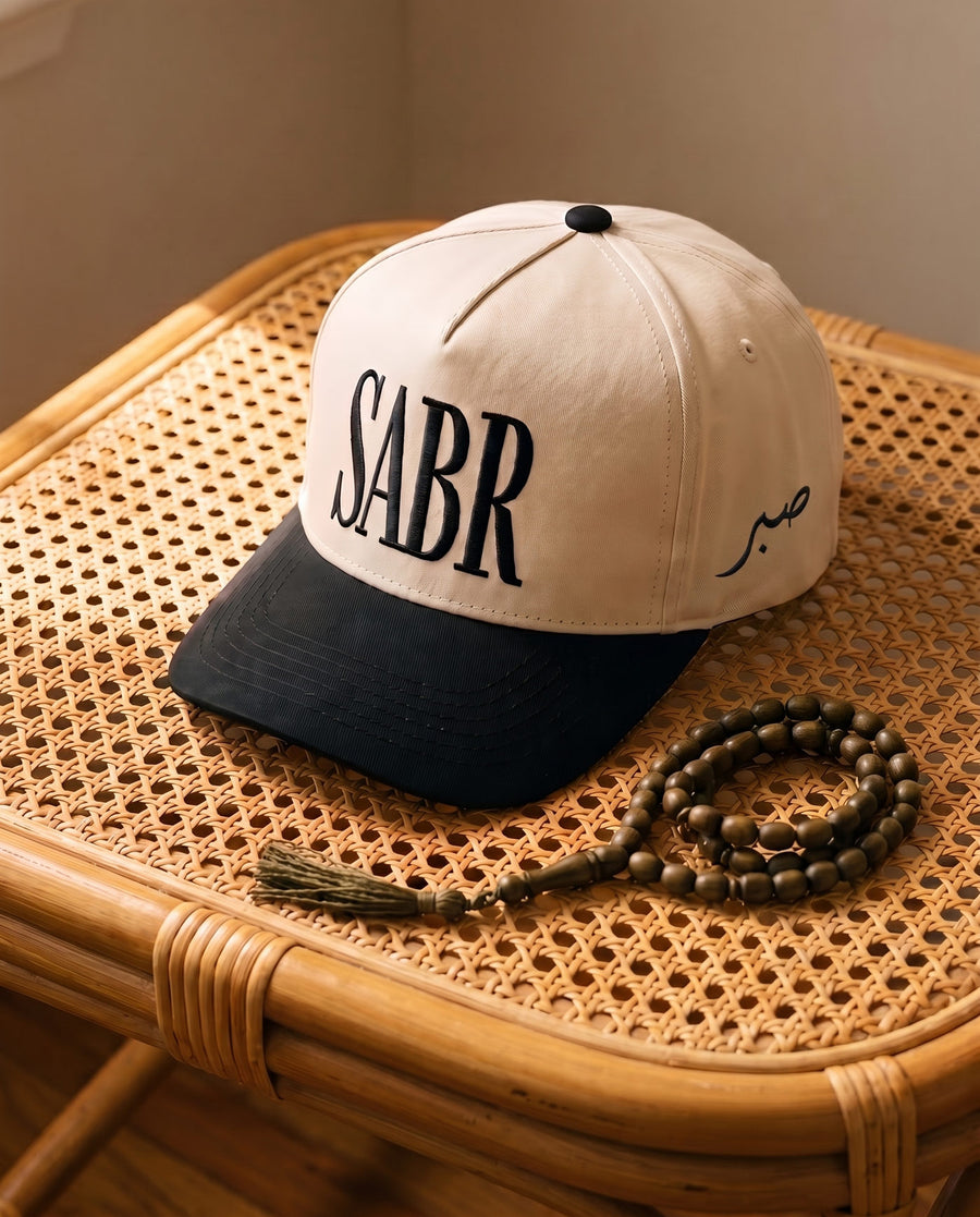 Sabr Cap in ivory with a black brim and black embroidered lettering, on a cane table beside wooden prayer beads