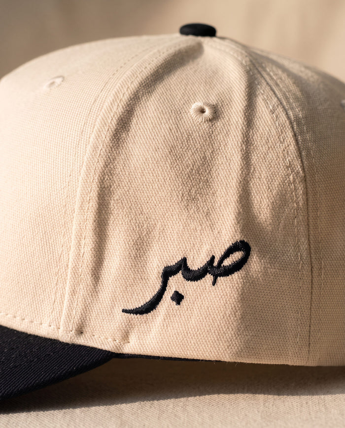 Close detail of the Arabic word sabr embroidered in black on the left side panel of the ivory Sabr Cap