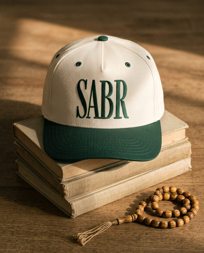 Sabr Cap in ivory with a dark green brim and dark green embroidered lettering, resting on a stack of vintage books beside a wooden misbaha