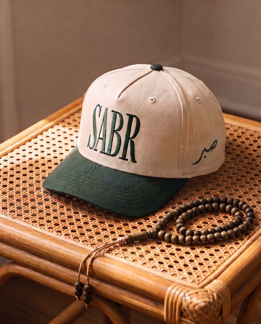 Sabr Cap in ivory with a dark green brim and dark green embroidered lettering, on a cane table beside wooden prayer beads