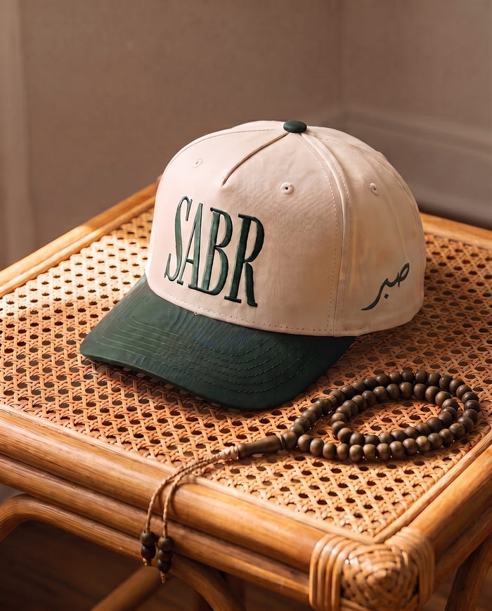 Sabr Cap in ivory with a dark green brim and dark green embroidered lettering, on a cane table beside wooden prayer beads