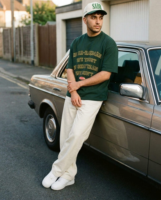 Sabr Cap in ivory with a dark green brim worn with the forest In Ar-Rahman We Trust tee, leaning on a car