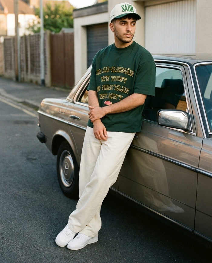 Sabr Cap in ivory with a dark green brim worn with the forest In Ar-Rahman We Trust tee, leaning on a car