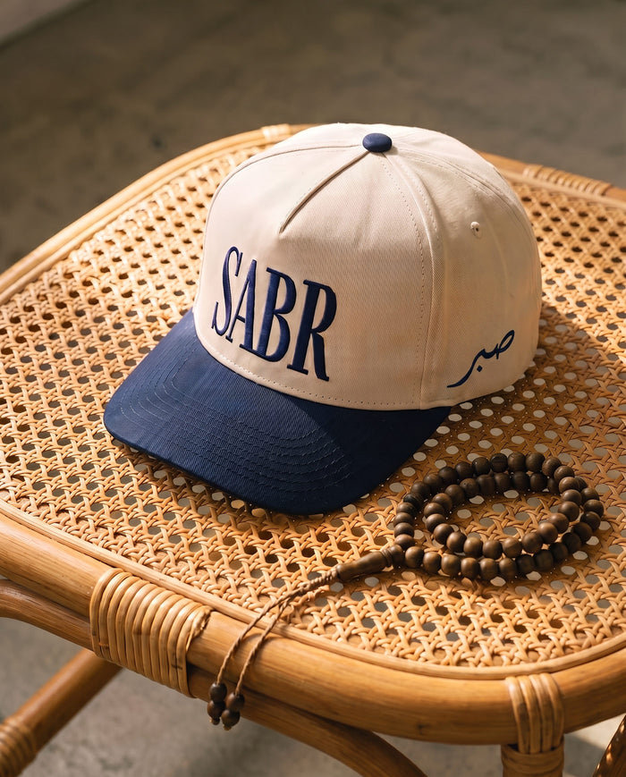 Sabr Cap in ivory with a navy brim and navy embroidered lettering, on a cane table beside wooden prayer beads