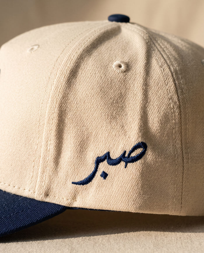 Close detail of the Arabic word sabr embroidered in navy on the left side panel of the ivory Sabr Cap