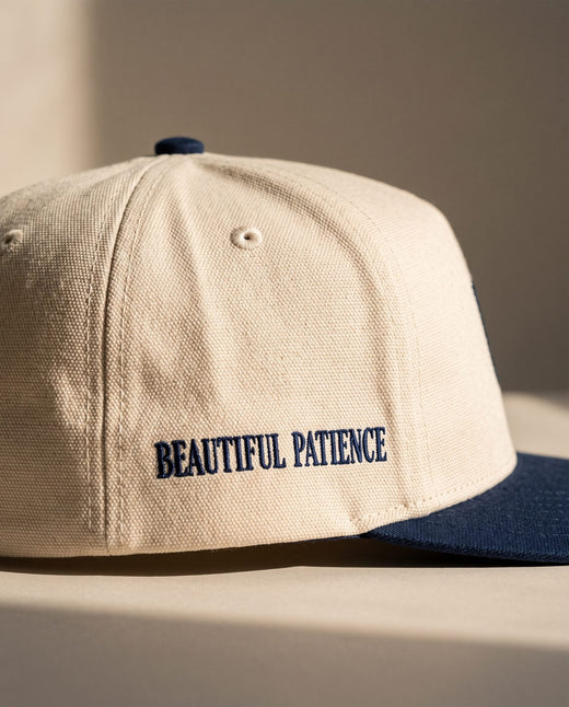 Close detail of BEAUTIFUL PATIENCE embroidered in navy on the right side panel of the ivory Sabr Cap
