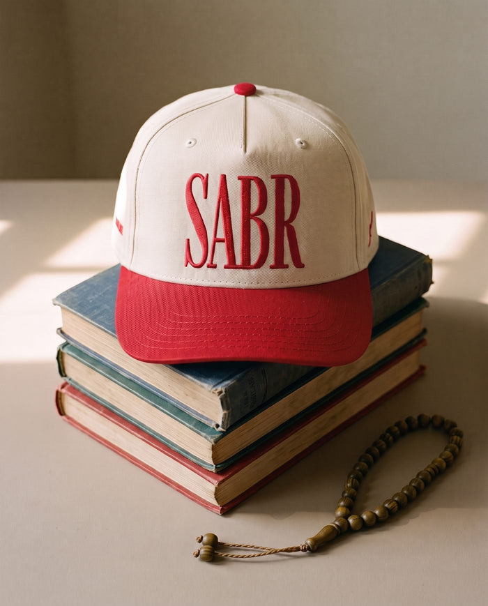 Sabr Cap in ivory with a red brim and red embroidered lettering, resting on a stack of vintage books beside a wooden misbaha