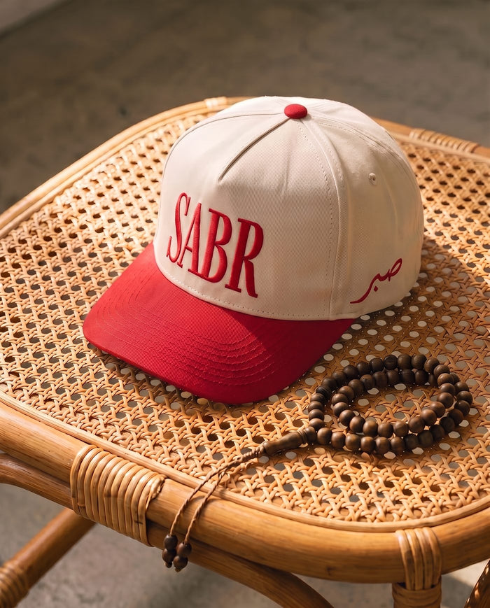 Sabr Cap in ivory with a red brim and red embroidered lettering, on a cane table beside wooden prayer beads