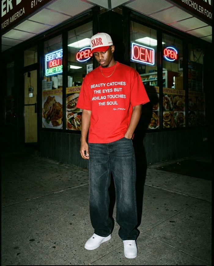 Sabr Cap in ivory with a red brim worn with the red Akhlaq Over Beauty tee outside a neon-lit deli at night
