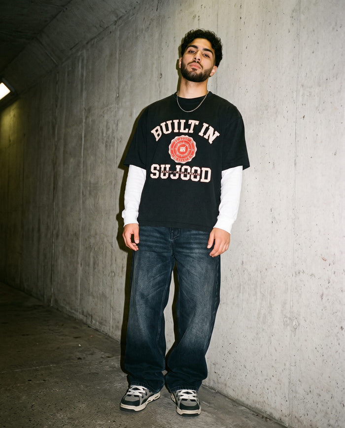 Built in Sujood black boxy tee over a white long sleeve against concrete