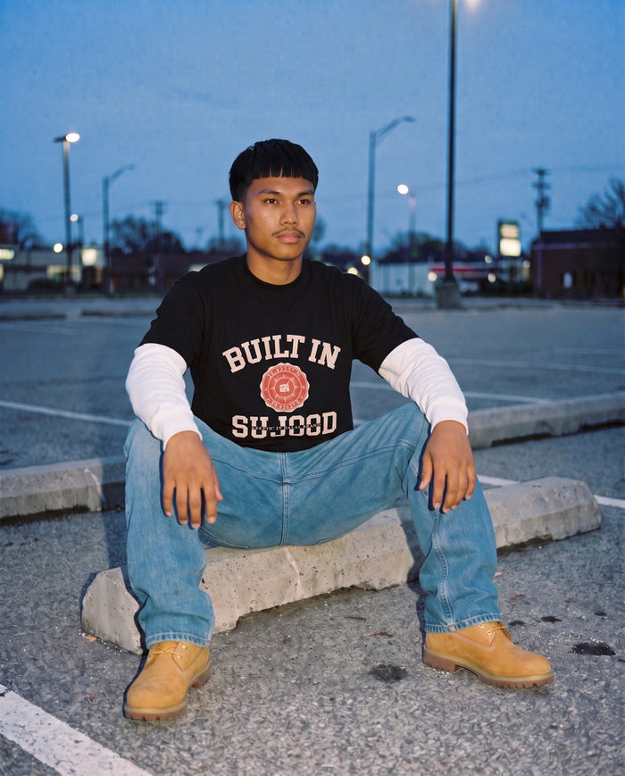 Built in Sujood black boxy tee, seated kerbside at dusk